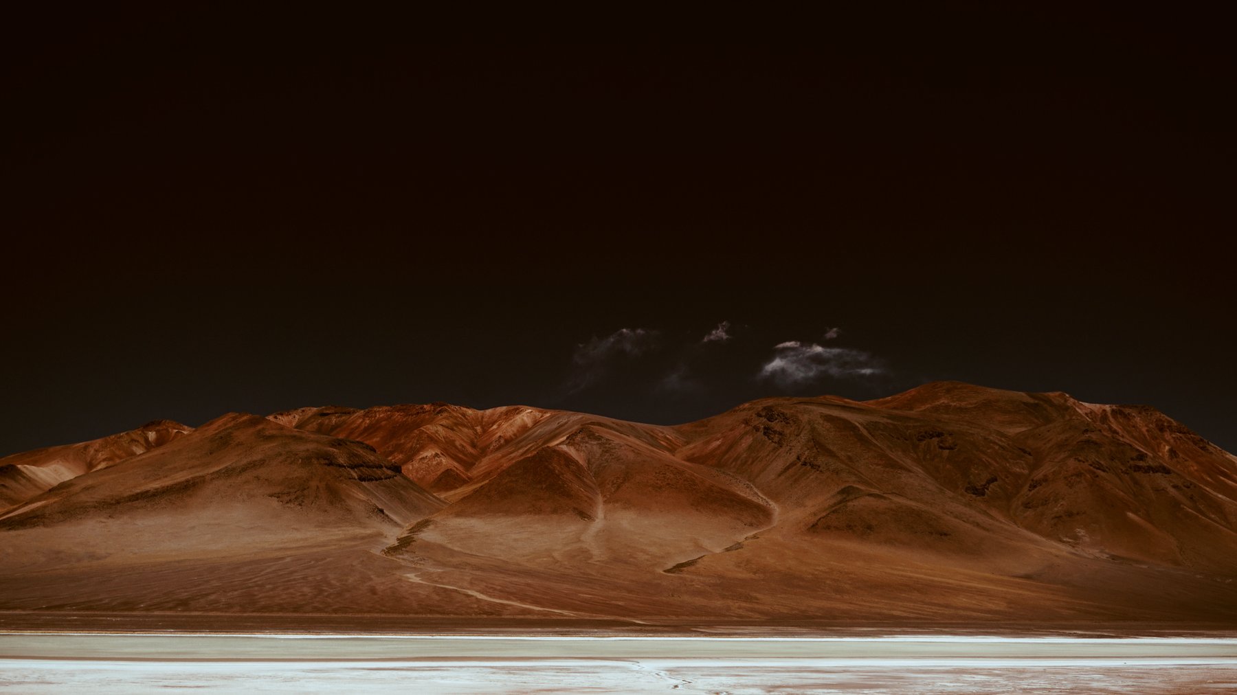 Reddish desert mountains beneath a dark sky above a salt flat